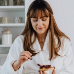 Woman in a bathrobe spreading jam on toast in a bright kitchen.