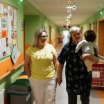 Two women walking in a nursing home corridor, one holding a child doll.